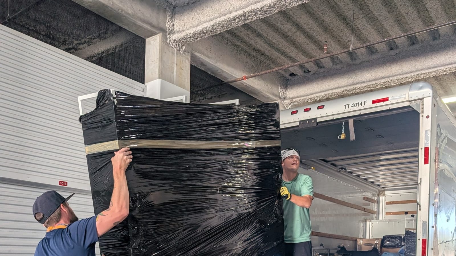 Two movers handling a large piece of furniture wrapped in black protective film near the truck ramp.